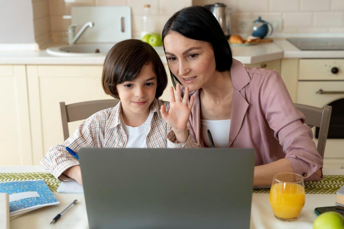 Mother and child waving at online tutor on laptop during home tutoring session at kitchen table